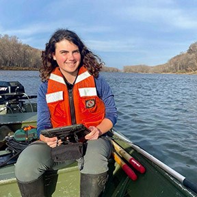 A girl with long black hair smiles at the camera while sitting on a green bench in a boat. The boat motor is visible behind her. She is wearing an orange life vest and holding a black electronic device with a cord that is running over the side of the boat