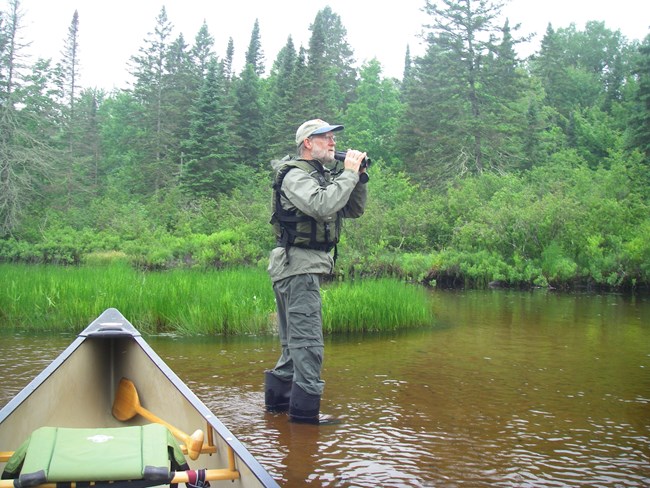 A man with binoculars stands in a river in front of his canoe