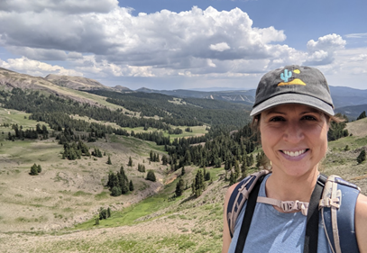 A woman wearing a ball cap stands on a high point overlooking broad valleys and conifer trees.