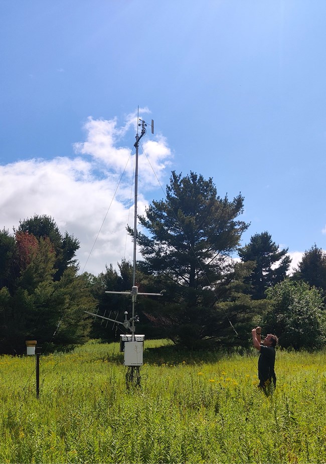 A man is looking up toward the top of a weather station in the middle of a open field.