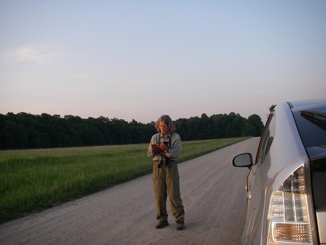 Photo of a woman standing in the middle of a gravel road beside her car. A field to her left extends behind her to a line of trees.