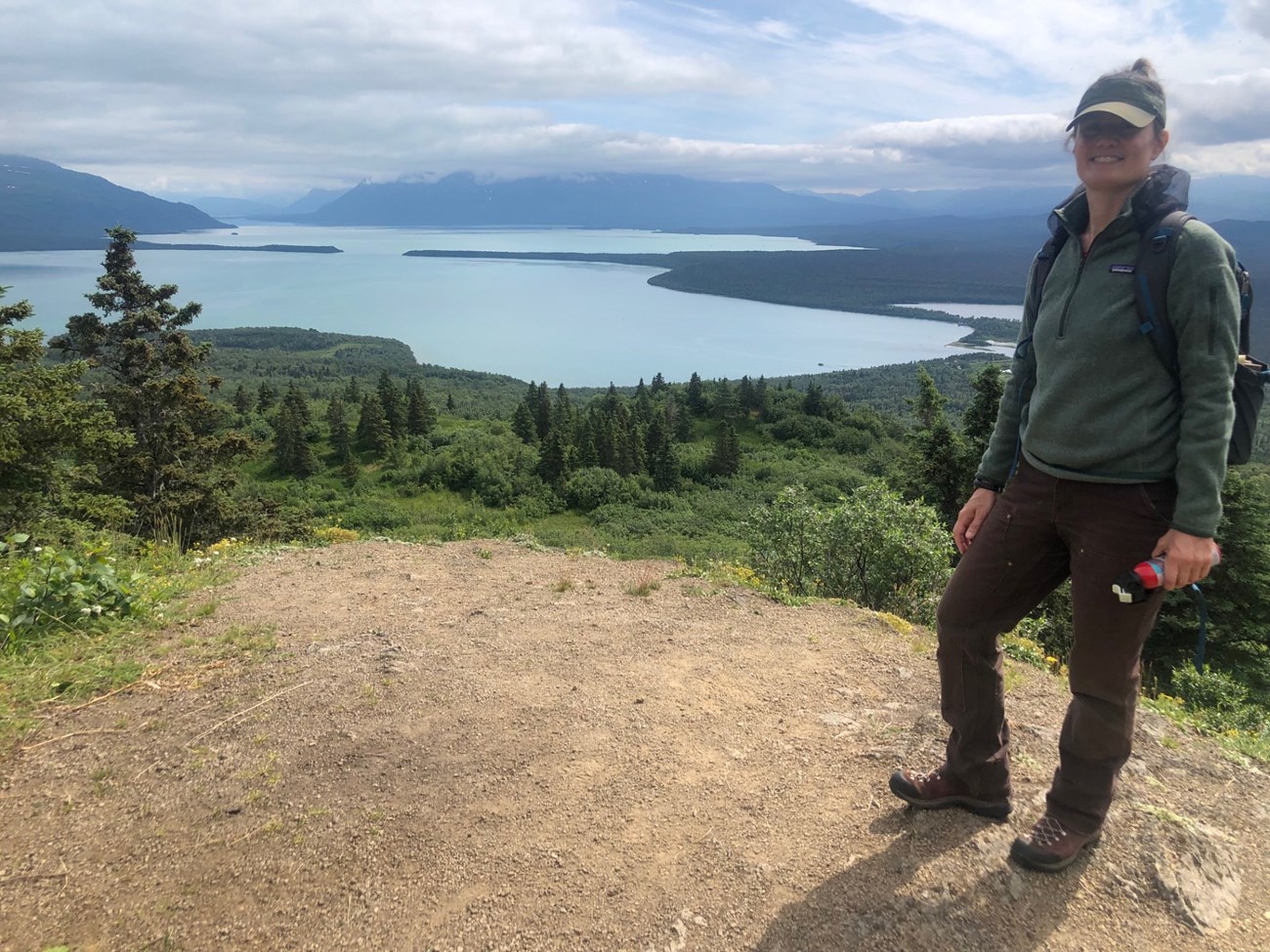 Woman wearing green jacket and brown pants stands on a gravelly overlook.