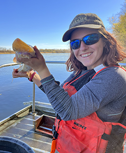 A person standing on a boat in a lifejacket holding a fish up to the camera.