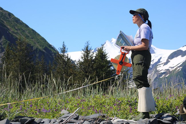 NPS employee holding a measuring tape and notes; snowy mountains in the background