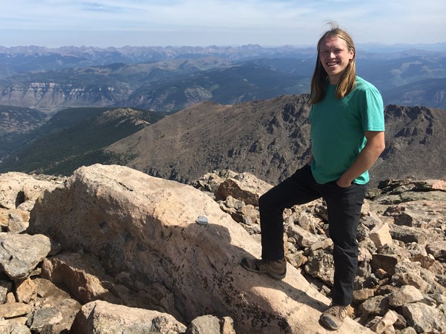 Man in green shirt standing on rocky summit with mountains in the distance