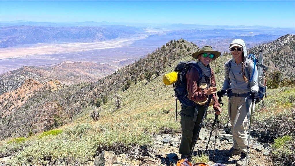 Two women in sun-protective clothing and backpacks full of gear standing on a sparsely vegetated ridge high above a desert valley floor.