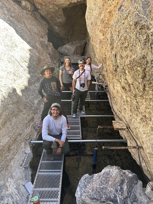Five people standing on metal grating suspended between two rock walls and spanning a chasm full of water.