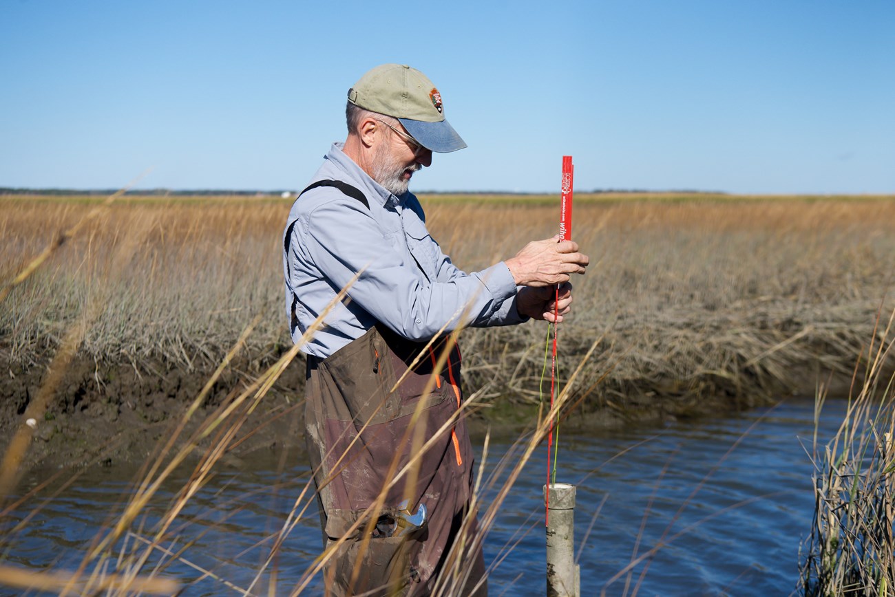 A field crew member stands holding a 6inch red rod over a pipe in a river