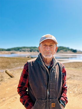 Scientist stands in front of riverbed wearing a hat and vest.