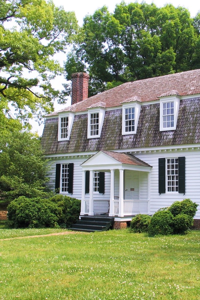A two-story white stately house with brown barn roof and chimney stands on a lawn with trees