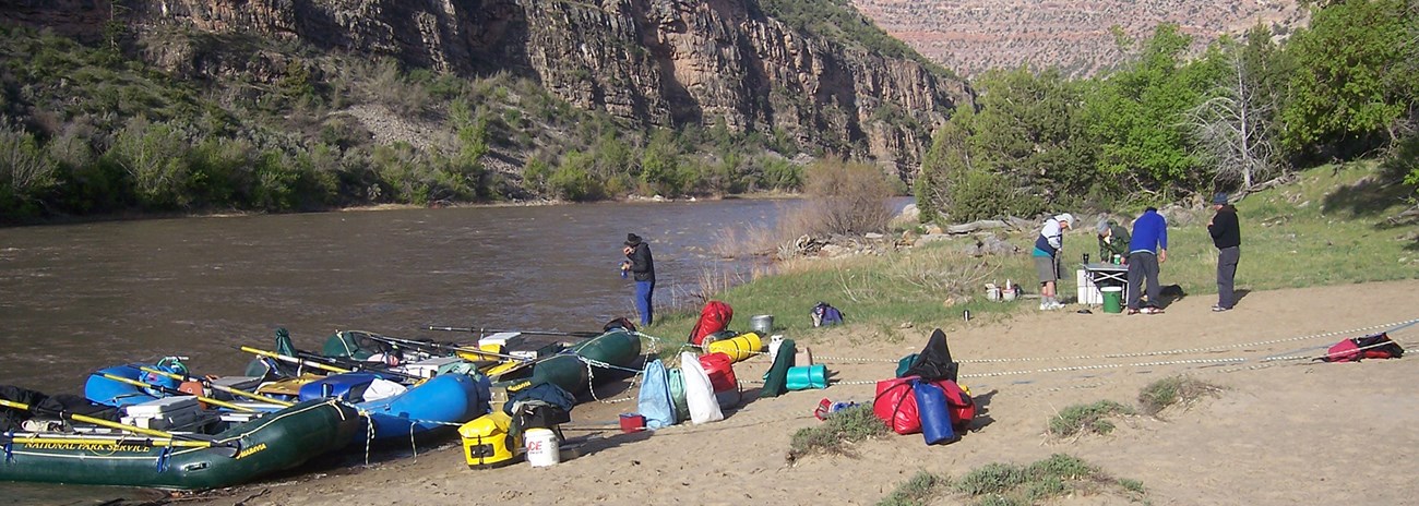 Several people stand around a stove at a river camp. The camp is sandy and open, with trees nearby and cliffs across the river. Four rafts are pulled up onshore.
