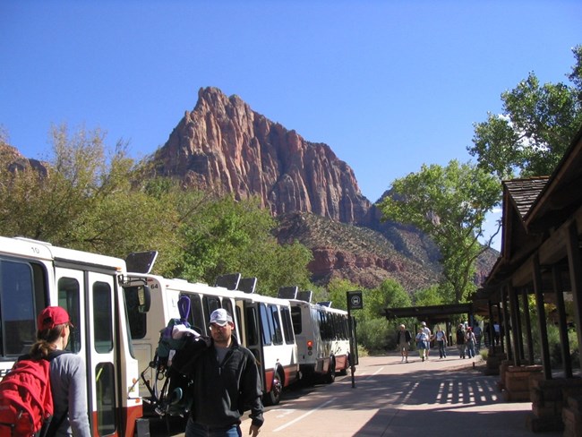 Visitors board the park shuttle at Zion’s Visitor Center beneath towering red cliffs.