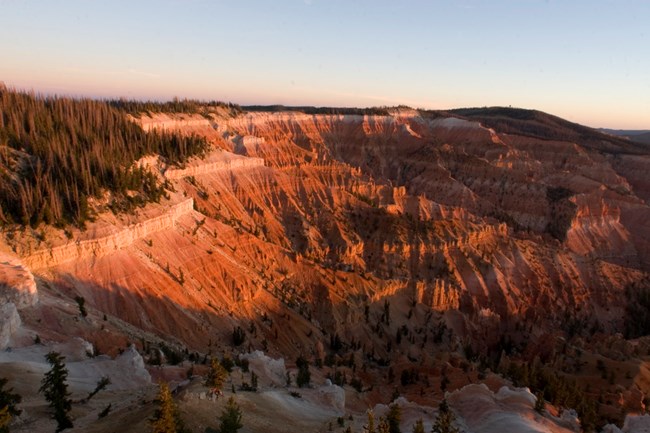 Cedar Breaks Amphitheater lit by sunset, revealing deep orange and red geologic formations.
