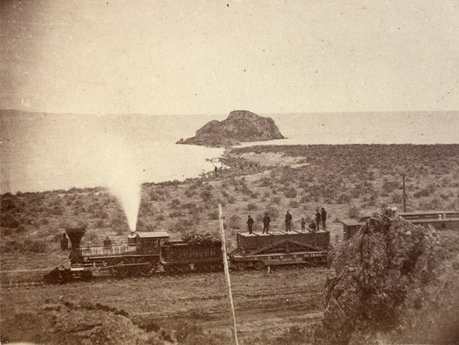 Steam locomotive passes near shoreline of Great Salt Lake with workers standing on flatcars