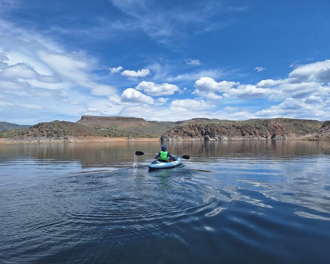 Kayaker paddles across calm waters of Blue Mesa Reservoir under a partly cloudy sky.