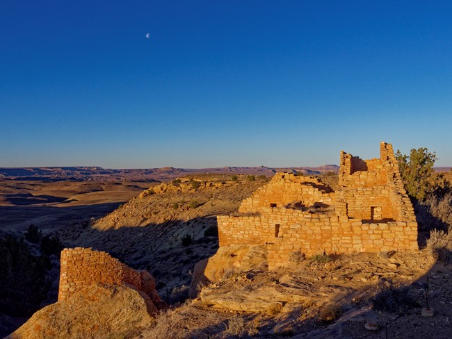 Crescent moon at sunset in a deep blue sky above a high desert landscape, with ancestral Puebloan sandstone structures in the foreground on the south end of Cajon Mesa.