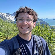 A young white man with curly brown hair and glasses smiles on a mountain top.