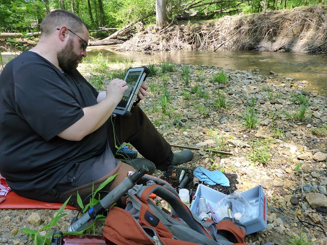 A man sits near a stream to enter data into a tablet computer.