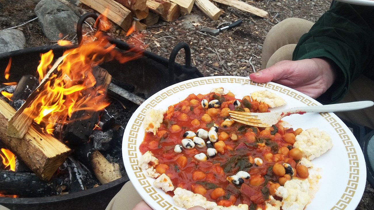 A hand holding a white plate with food and a fork on it and a campfire in the background.
