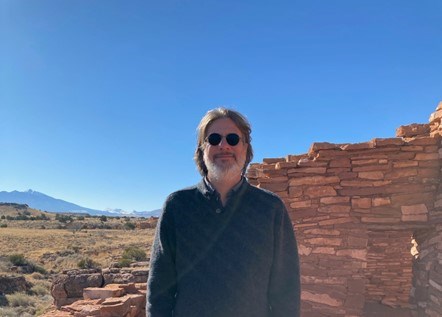 Man standing in front of ancestral Puebloan structure on a clear, blue-sky day.