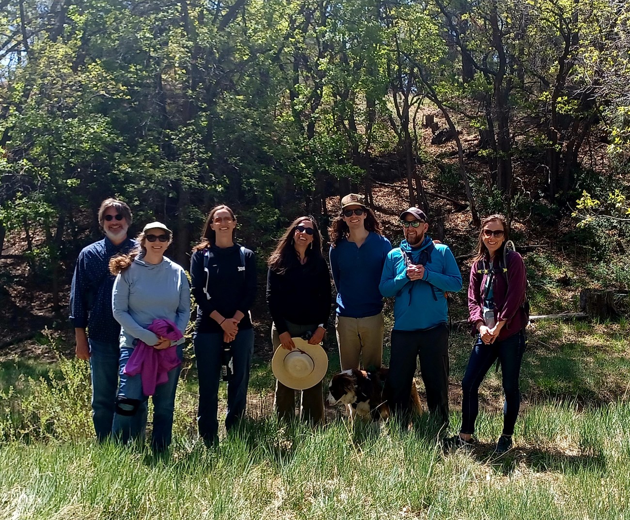 Seven smiling people and one dog standing side by side in an oak forest.