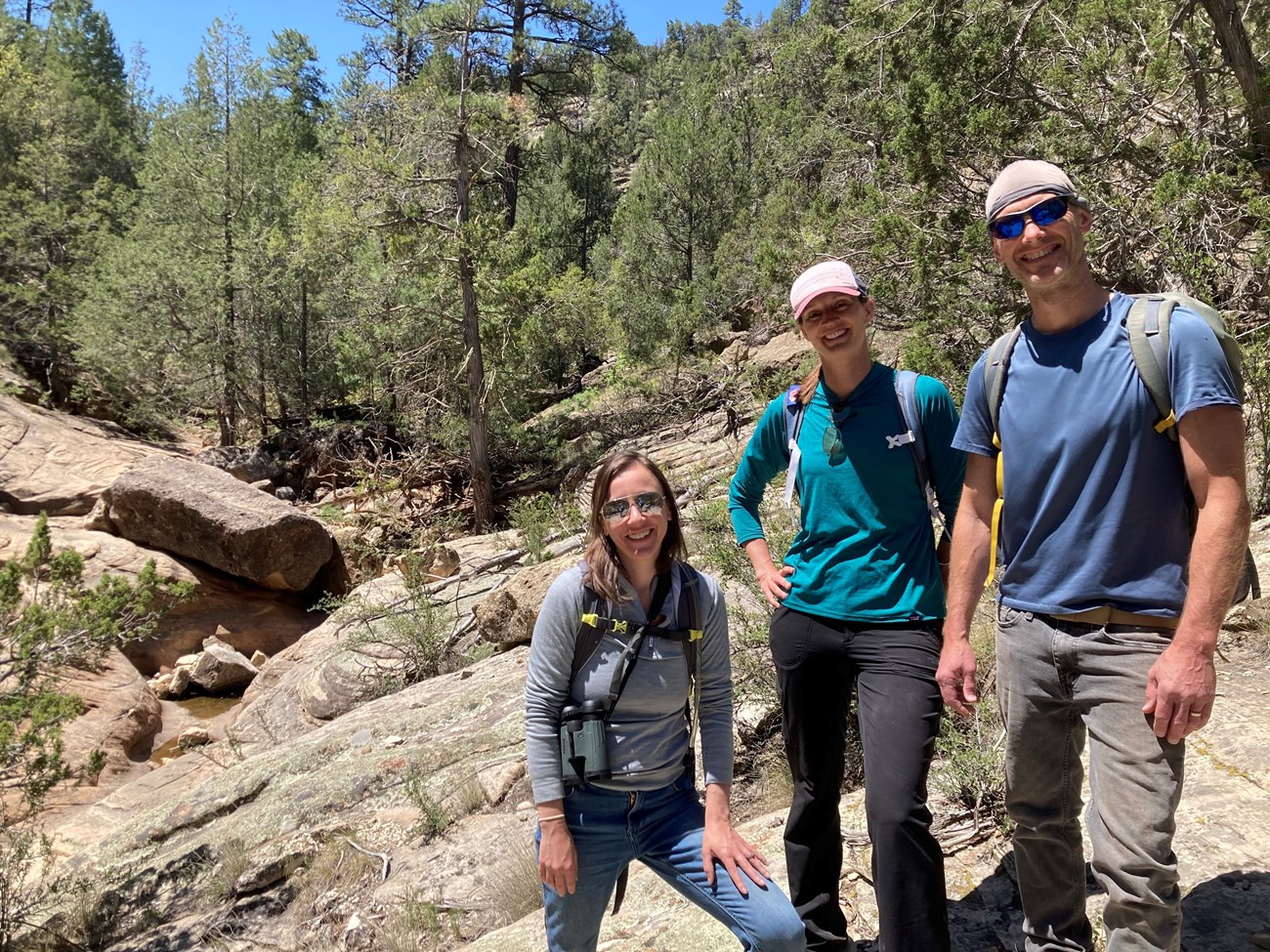 Three people standing together and smiling on a large rock in the forest.