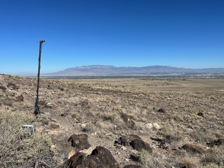 A long black pole mounted in the desert with a recording device on type.