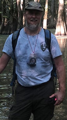 Man with hat and backpack standing in water under cypress trees