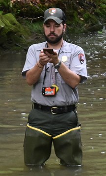 Man in uniform wearing waders in a stream holding a device