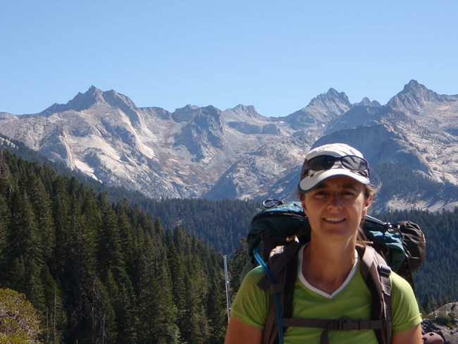 Woman with a backpack smiles at camera with a views of conifer forest and a rugged mountain range.