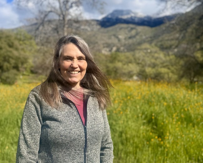 A woman smiles in front of a bright green meadow with yellow flowers. A few trees just starting to leaf out and a mountain is visible in the background.