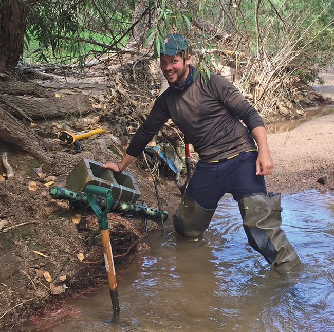 Photo of man in hip waders standing in a stream. He leans on a concrete block sitting on the bank. A long tube with holes in it, used for sonde deployment, is attached to the block. A shovel stands in the water.