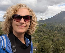 A woman with curly blond hair and sunglasses in front of a volcano surrounded by green trees.