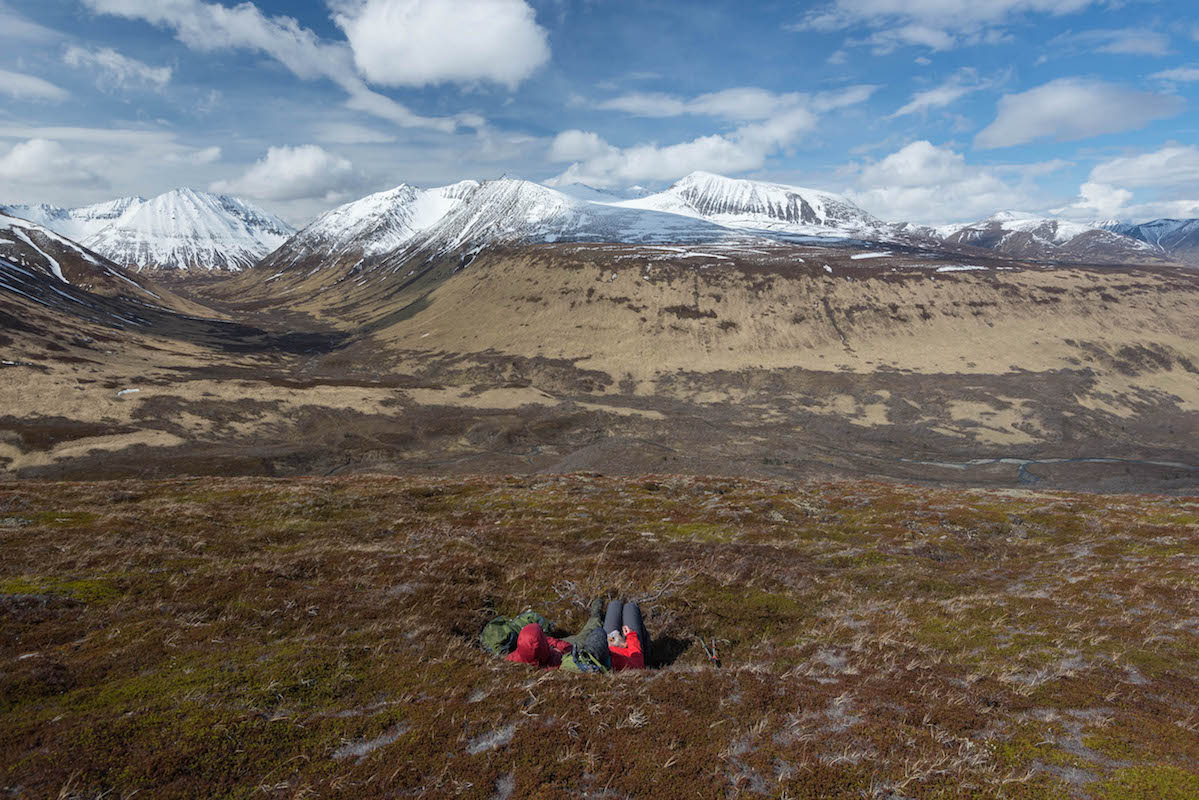 Two people lay on the tundra looking out at mountains and a river valley