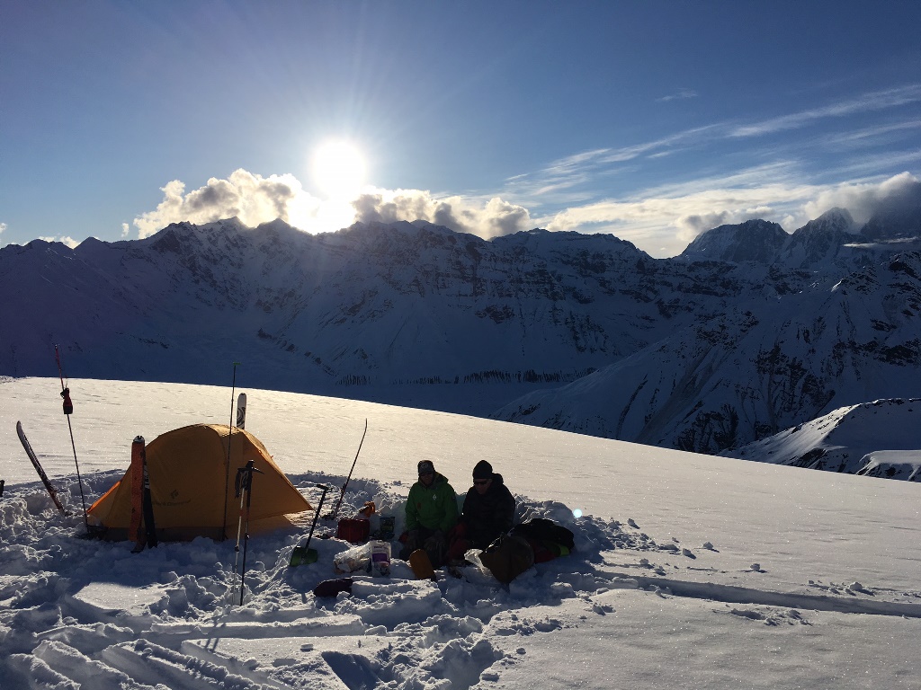 Two climbers sit outside a tent on a snowfield overlooking distant mountain peaks