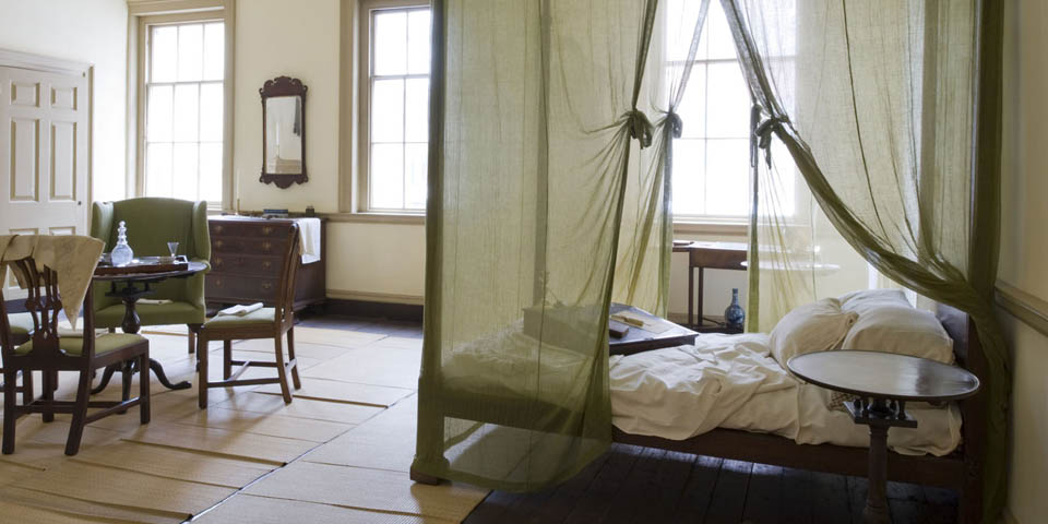 Color photo of the Bishop's bedroom with a four poster bed draped with thin gauze mosquito netting in the foreground and a table surrounded by mahogany chairs and an easy chair in the background.