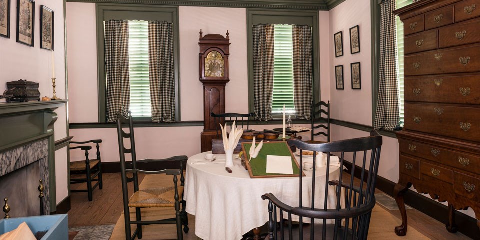 Color photo of the Declaration House parlor with a Windsor chair and table, tall chest, and tall case clock.