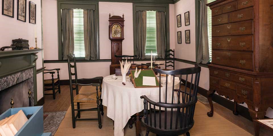 Color photo showing parlor with Windsor chairs around table in foreground, and tall case clock on pier wall in background.