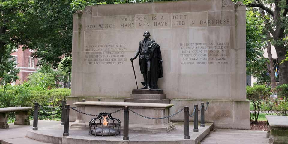 Color photo of statue of George Washington on plinth in front of tomb and eternal flame.  Behind Washington is a wall with text reading "Freedom is a light for which many men have died in darkness."