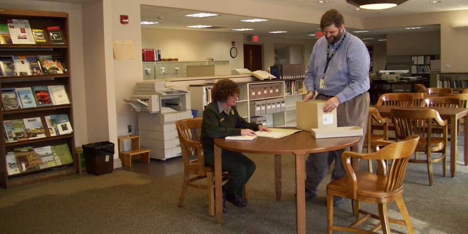 Color photo of an archivist putting a box of archival material on a table near a seated female park ranger.