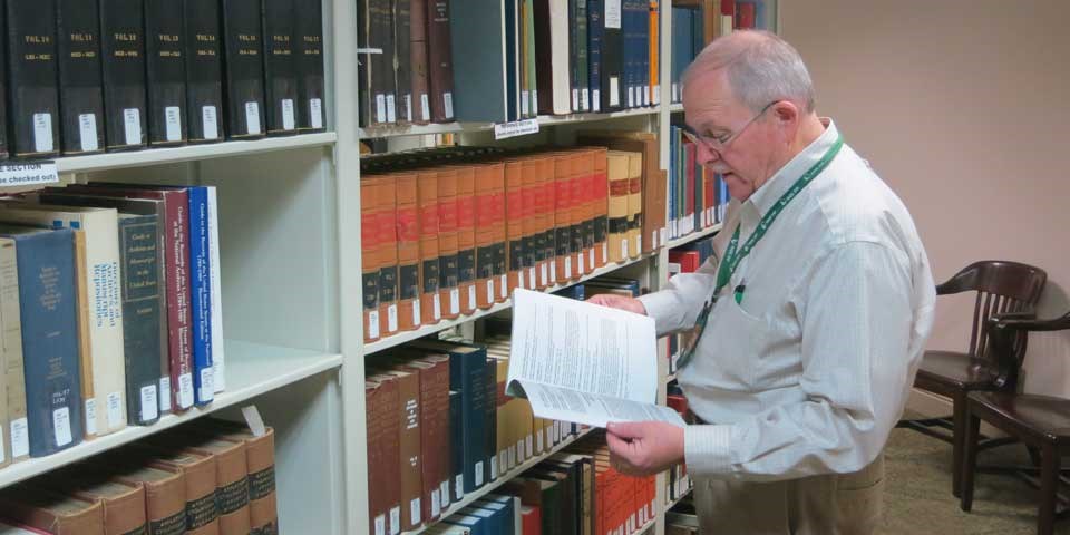 Color photo of a man standing next to a bookshelf in a library, holding a book.