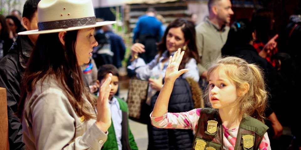 Color photo of female park ranger swearing in young girl as junior ranger.