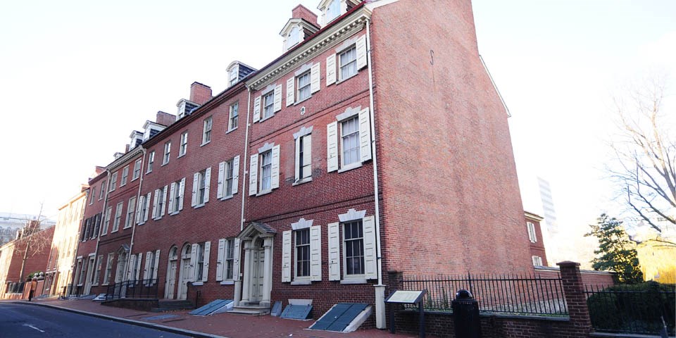 Color photo of a row of red brick rowhouses with buff colored shutters. The Bishop White House is the first house on the right and has a doorway with columns and triangular pediment.