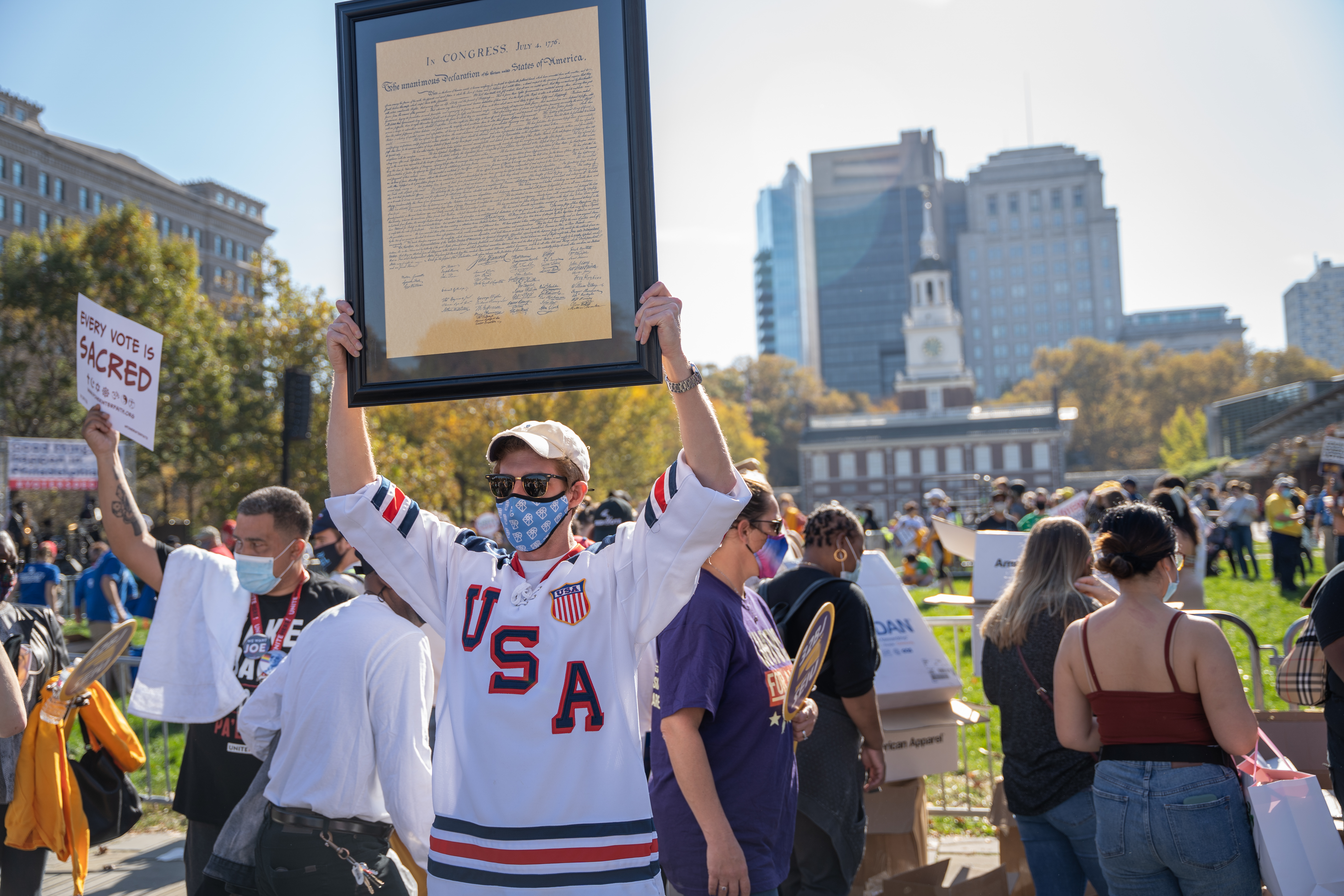 A man in the foreground wearing a USA jersey, sunglasses, and a mask holds a copy of the Declaration of Independence in a frame. Behind the man, people are gathered with signs near Independence Hall. Blue sky and cityscape in the background.