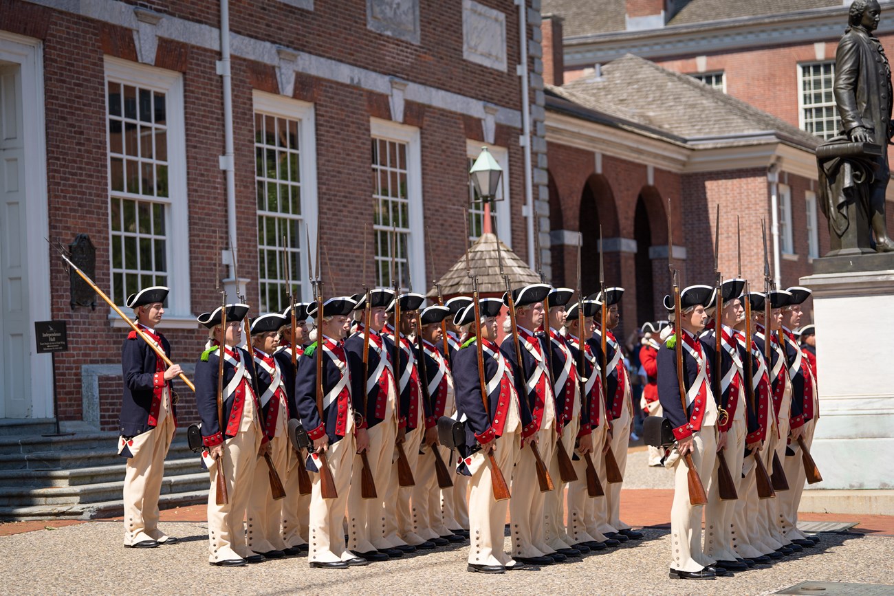 People in colonial military uniform stand in formation in front of a brick building