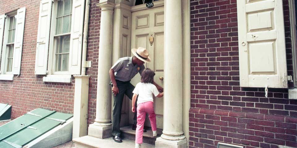 Color photo of a male park ranger and a small child  standing next to each other on a doorstep, working together to turn the doorknob.