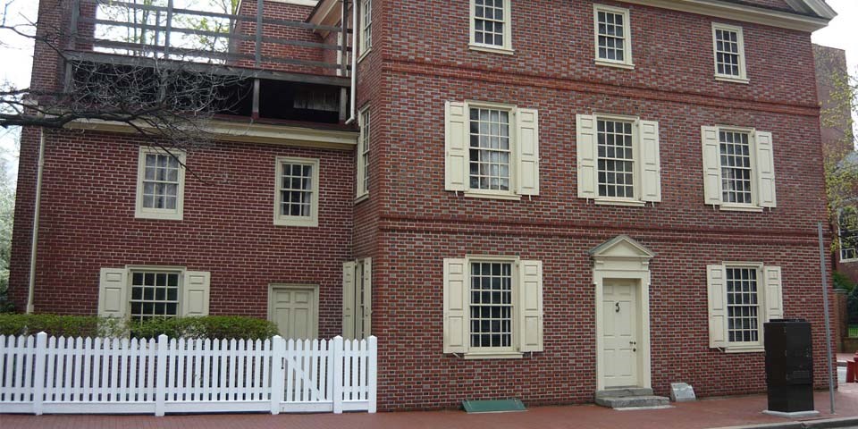 Color photo of two attached red brick buildings with a white fence in front of the smaller building.