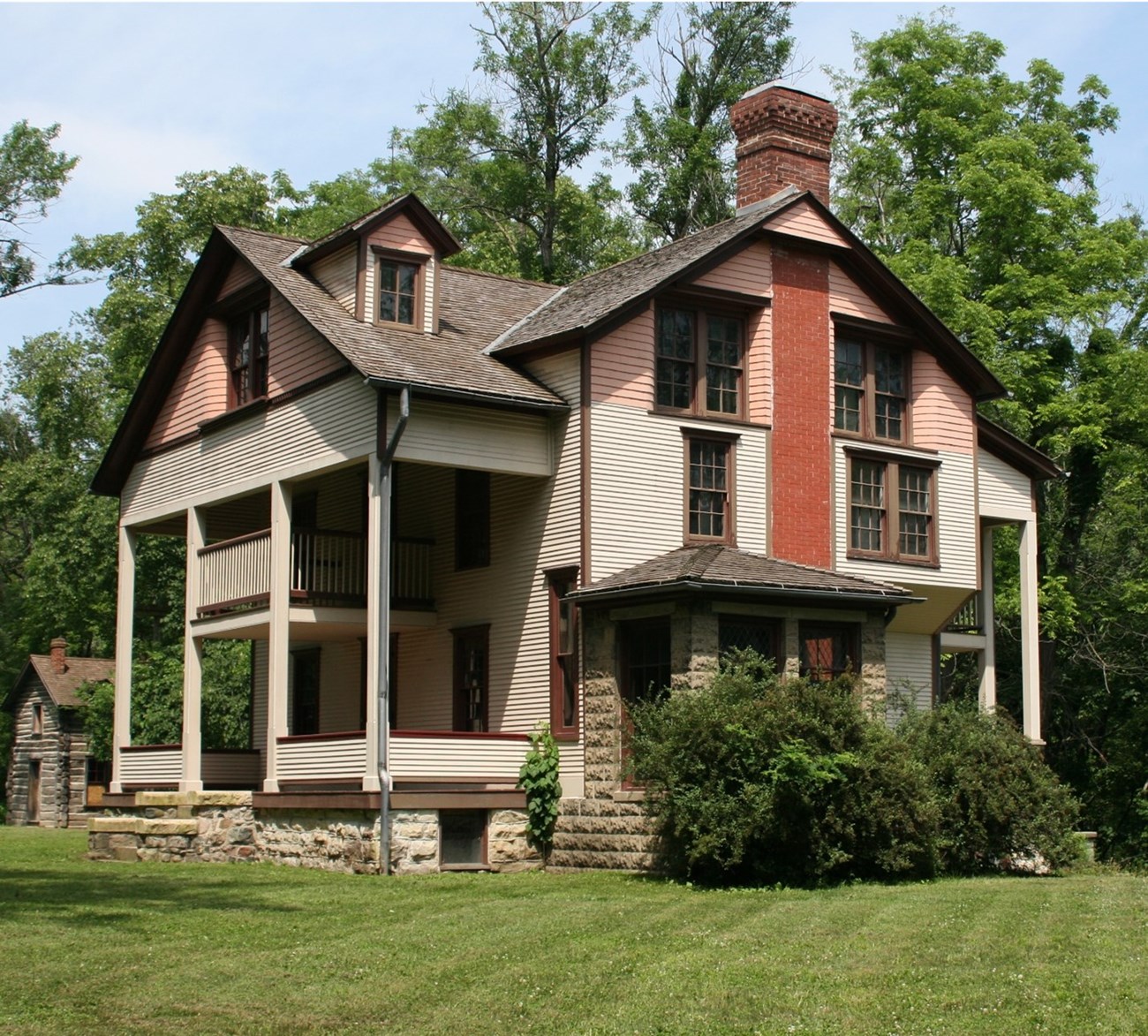 The multi-story Bailly Homestead sits on a manicured lawn.  The main house has 3 stories and features multi-colored horizontal siding along the upper structure and gray colored stone work along the foundation.