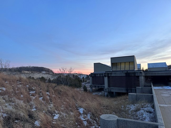 Sunset scene looking south east with the West Beach Bath House standing beside tawny dune grasses beneath a blue sky with hazy pink near the horizon.