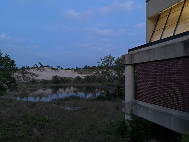 Photograph of the edge of the concrete West Beach Bath House, next to a wetland pond. The gray-blue sky of an expired day above.